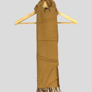 Könnte beinhalten: Ein beigefarbener Schal mit Fransen hängt an einem Holzbügel. Der Schal ist einfarbig mit einer strukturierten Optik und einem gefalteten Design. Der Schal ist aus einem weichen Material gefertigt.