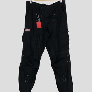 Puede incluir: Pantalones cargo negros con múltiples bolsillos y detalles de anillos metálicos plateados. Incluye un parche con la bandera estadounidense y una etiqueta roja. Los pantalones cuelgan de una percha de madera.