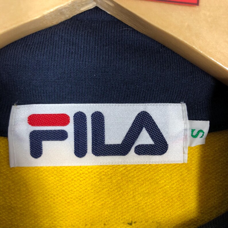 May include: A white tag with the word "FILA" in blue and red lettering on a navy blue and yellow polo shirt. The tag also has a size "S" in green lettering.