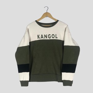 Może przedstawiać: Bluza z okrągłym dekoltem z napisem "KANGOL" w czarnych literach. Bluza ma wz&oacute;r w bloki kolor&oacute;w z białymi, oliwkowymi i czarnymi paskami. Odzież wisi na drewnianym wieszaku.