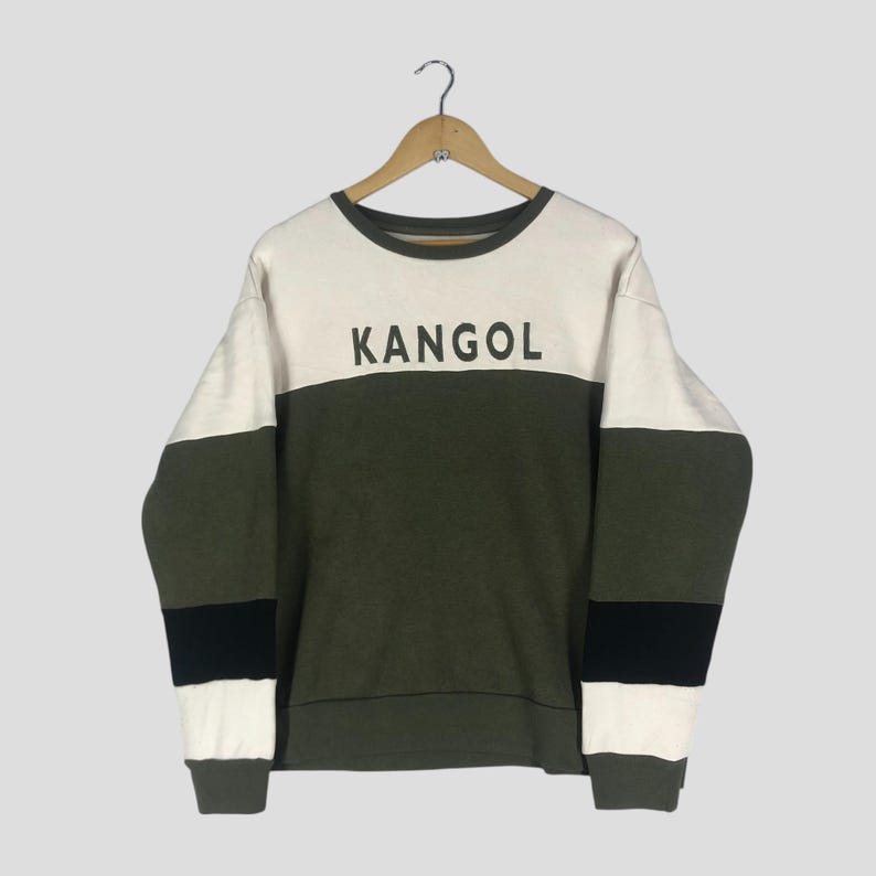 Może przedstawiać: Bluza z okrągłym dekoltem z napisem "KANGOL" w czarnych literach. Bluza ma wz&oacute;r w bloki kolor&oacute;w z białymi, oliwkowymi i czarnymi paskami. Odzież wisi na drewnianym wieszaku.