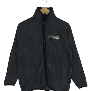 Può includere: Una giacca con zip in pile grigia con una cerniera nera e una piccola patch con logo sul petto sinistro che recita "Penny's"