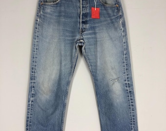 May include: Faded light blue denim jeans with a button fly, hanging on a wooden hanger. The jeans have a distressed appearance with wear and a red tag attached. The jeans have a worn look.