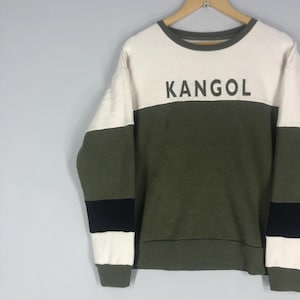 Może przedstawiać: Bluza w blokach kolor&oacute;w z napisem "KANGOL" w czarnych literach. G&oacute;rna część jest w kolorze złamanej bieli, środkowa część jest oliwkowozielona, a rękawy mają czarno-białe paski. Bluza wisi na drewnianym wieszaku.