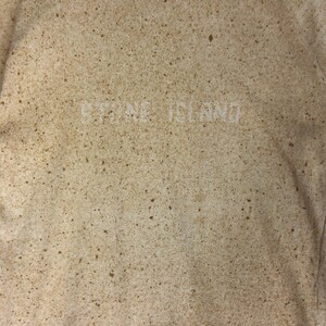 May include: A beige sweatshirt with a speckled pattern and the text "STONE ISLAND" in white. The neckline is a lighter beige color. The overall design is a neutral, textured look.