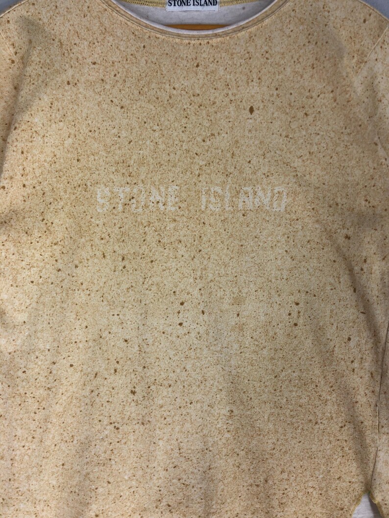 May include: A beige sweatshirt with a speckled pattern and the text "STONE ISLAND" in white. The neckline is a lighter beige color. The overall design is a neutral, textured look.