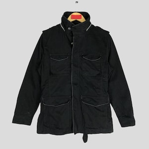 May include: Black jacket with four front pockets and a high collar. The jacket has a button closure and a small loop detail at the waist. The jacket is hanging on a wooden hanger.