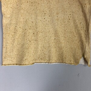 May include: A tan-colored t-shirt with a speckled pattern and the text "STONE ISLAND" printed across the top. The shirt has a raw hemline and a white undershirt peeking out from the bottom.