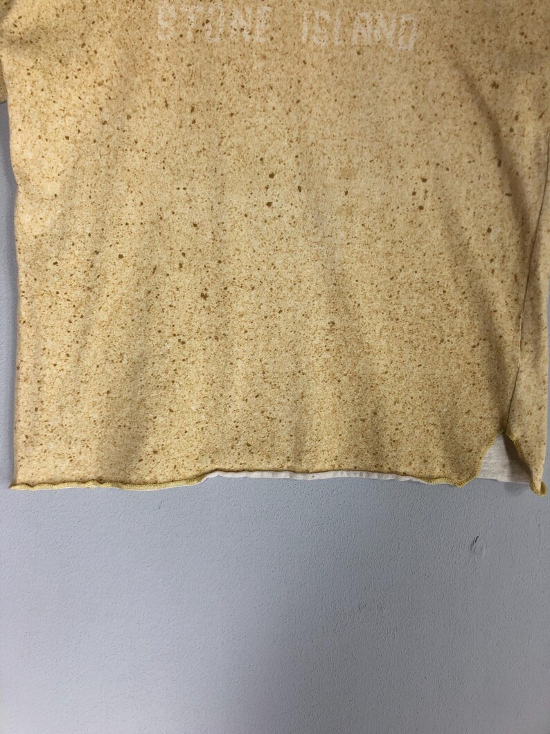 May include: A tan-colored t-shirt with a speckled pattern and the text "STONE ISLAND" printed across the top. The shirt has a raw hemline and a white undershirt peeking out from the bottom.
