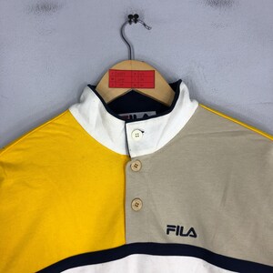 May include: A multi-coloured polo shirt with a high collar and button closure. The shirt is yellow, beige, and white with a navy blue stripe at the bottom. The shirt has the word "FILA" embroidered on the front in navy blue.