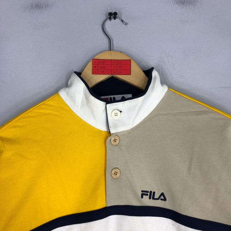 May include: A multi-coloured polo shirt with a high collar and button closure. The shirt is yellow, beige, and white with a navy blue stripe at the bottom. The shirt has the word "FILA" embroidered on the front in navy blue.