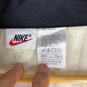May include: White Nike windbreaker jacket with a black zipper and black trim. The jacket has a white label with the Nike logo and care instructions. The label also includes the size, L, and the text "Made in Thailand, RN #56323."