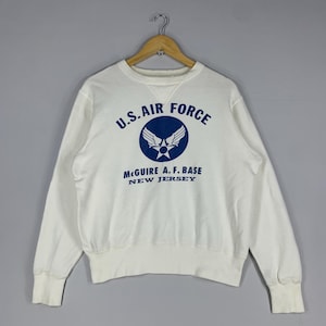 May include: A white crew neck sweatshirt featuring the text "U.S. AIR FORCE" in blue, a circular emblem, and the text "McGuire A.F. BASE NEW JERSEY". The sweatshirt has ribbed cuffs and hem.