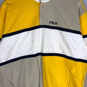 May include: A long-sleeved shirt in yellow, white and beige with a black stripe across the chest. The shirt has a button-up collar and the word "FILA" is printed on the chest.