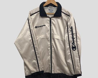 May include: Cream-colored Champion products jacket with black trim and a black collar. The jacket features the Champion logo on the chest and sleeve. The jacket is hanging on a wooden hanger.