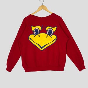 May include: Red crewneck sweatshirt featuring a large yellow and blue cartoon bird face on the front. The bird has blue eyes and a yellow beak. The sweatshirt is hanging on a wooden hanger.