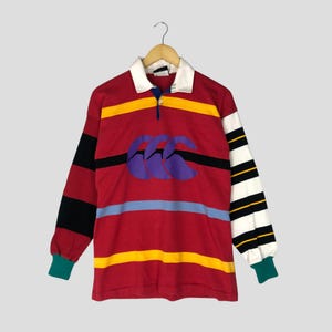 May include: A red rugby shirt with horizontal yellow, black, and blue stripes. The long sleeves are striped black and white on one side and solid black on the other. The shirt features a white collar and the Canterbury logo.