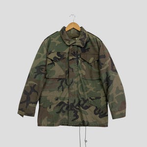 May include: A long-sleeve field jacket featuring a woodland camouflage pattern in shades of green, brown, and black. The jacket includes a fold-down collar, two button-flap chest pockets, two button-flap waist pockets, and a front zip fastening.