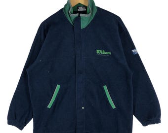 Könnte beinhalten: Marineblaue Wilson Fleecejacke mit grünen Akzenten. Mit hohem Kragen, zwei Vordertaschen und dem grün gestickten Wilson-Logo auf der Brust. Eine Jacke im Vintage-Stil.