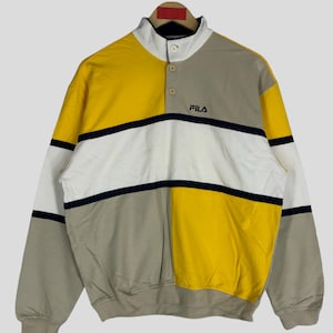 May include: A yellow, white and grey striped sweatshirt with a navy blue collar and a button placket. The sweatshirt has the word "FILA" in navy blue lettering on the front.