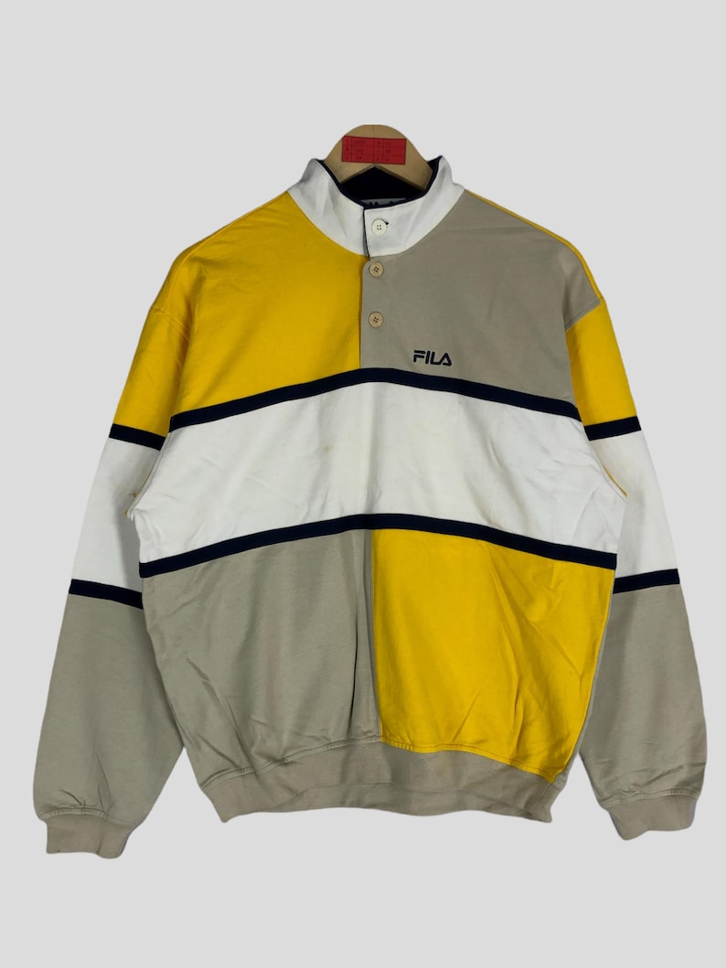 May include: A yellow, white and grey striped sweatshirt with a navy blue collar and a button placket. The sweatshirt has the word "FILA" in navy blue lettering on the front.