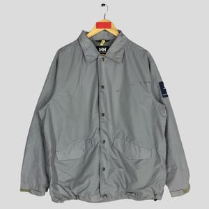 May include: A gray, long-sleeved jacket with a button-up front and a drawstring at the waist. The jacket has a patch on the left sleeve with the text "Helly Hansen" in white.