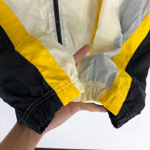 May include: Close-up of a black, yellow, and white windbreaker jacket. The jacket has a zipper closure and a drawstring hood. The jacket is made of a lightweight, water-resistant material.