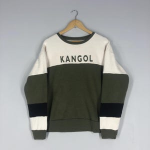 Może przedstawiać: Bluza z okrągłym dekoltem z napisem "KANGOL" w czarnych literach. G&oacute;rna część jest biała, środek oliwkowy, a rękawy mają oliwkowo-zielone i czarne paski. Bluza wisi na drewnianym wieszaku.