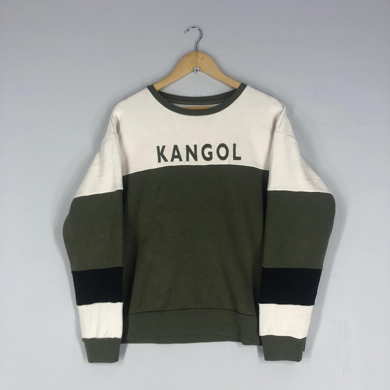 Może przedstawiać: Bluza z okrągłym dekoltem z napisem "KANGOL" w czarnych literach. G&oacute;rna część jest biała, środek oliwkowy, a rękawy mają oliwkowo-zielone i czarne paski. Bluza wisi na drewnianym wieszaku.