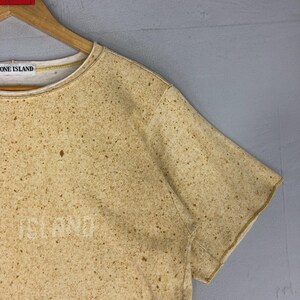 May include: Tan short-sleeved t-shirt with a speckled pattern. The shirt has the word "ISLAND" printed on the front and the words "STONE ISLAND" on the collar. The shirt is hanging on a wooden hanger.
