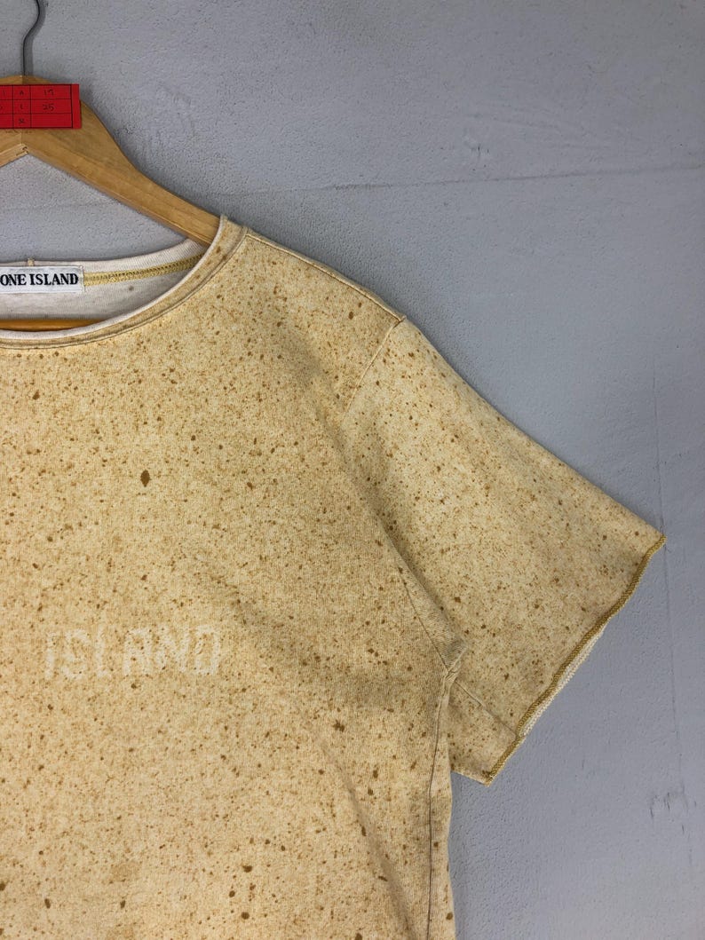 May include: Tan short-sleeved t-shirt with a speckled pattern. The shirt has the word "ISLAND" printed on the front and the words "STONE ISLAND" on the collar. The shirt is hanging on a wooden hanger.