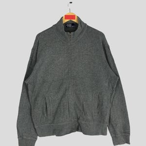 May include: A gray zip-up sweatshirt with a stand-up collar. The sweatshirt has a small logo on the left chest and a larger logo on the bottom right.