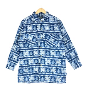 Puede incluir: Camisa de manga larga azul y blanca con estampado de copos de nieve y hojas. La camisa tiene cuello, dos bolsillos en el pecho y está abotonada. Cuelga de una percha de madera. La tela parece suave.