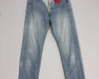 May include: Pale blue denim jeans with a faded effect, button fly, and a red tag. The jeans are suspended on a wooden hanger. The jeans have a classic, straight-leg fit. The background is plain.