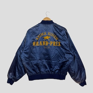 May include: A navy blue satin bomber jacket featuring gold embroidered text and a horse graphic. The text reads "ARIMA KINEN GRAND-PRIX" and "SINCE 1956". The jacket has a ribbed collar, cuffs, and hem in a darker navy blue.