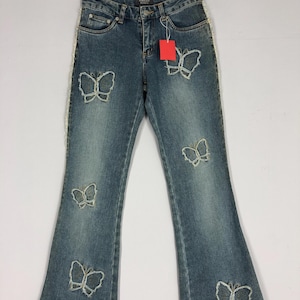 May include: Blue denim jeans with a flared leg design. The jeans feature butterfly cutouts and a white trim along the side seams. A red tag hangs from the waistband. The jeans are displayed on a hanger.
