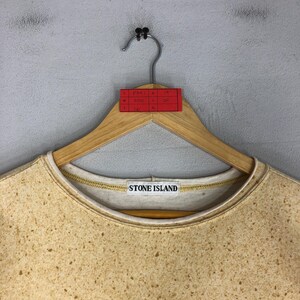 May include: A tan Stone Island sweatshirt with a speckled pattern, hanging on a wooden hanger. The sweatshirt has a white collar and the Stone Island logo is visible. A red tag with handwritten measurements is attached to the hanger.