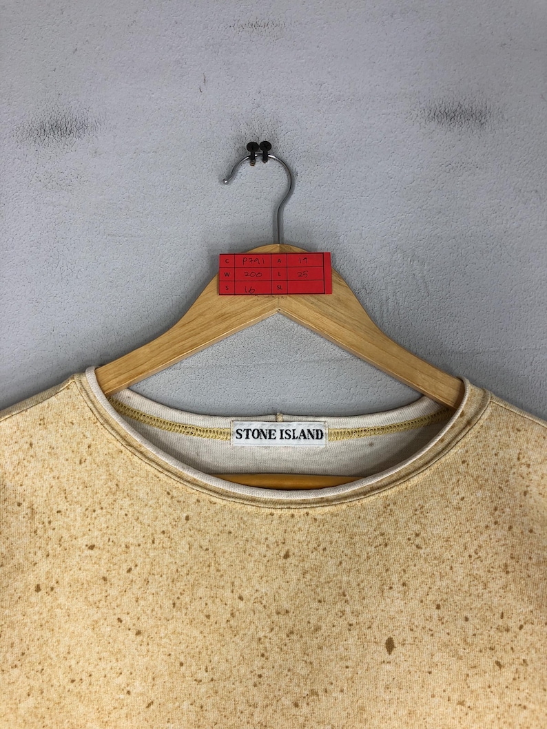 May include: A tan Stone Island sweatshirt with a speckled pattern, hanging on a wooden hanger. The sweatshirt has a white collar and the Stone Island logo is visible. A red tag with handwritten measurements is attached to the hanger.