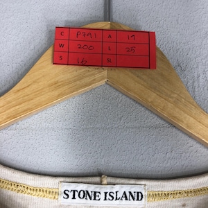 May include: A beige Stone Island shirt is displayed on a wooden hanger. A red tag with handwritten measurements and codes is attached to the hanger. The Stone Island logo is visible on the shirt's neckline.