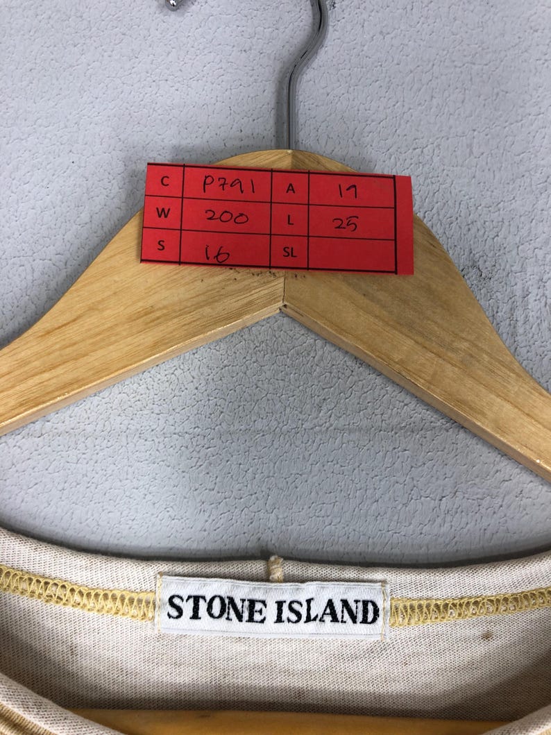 May include: A beige Stone Island shirt is displayed on a wooden hanger. A red tag with handwritten measurements and codes is attached to the hanger. The Stone Island logo is visible on the shirt's neckline.