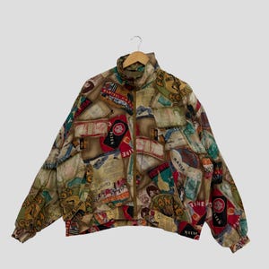 Könnte beinhalten: Eine Vintage-Jacke mit Reißverschluss und einem einzigartigen Collage-Muster. Die Jacke hat einen hohen Kragen, elastische Bündchen und eine braune und beige Farbgebung. Das Design integriert verschiedene Etiketten und Textelemente und erzeugt eine Retro-Ästhetik.