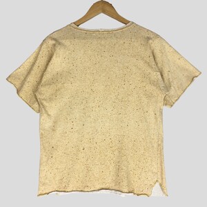 May include: A short-sleeved, beige t-shirt with a speckled pattern, hanging on a wooden hanger. The shirt has a raw hemline and a slightly distressed appearance. The fabric appears to be a textured cotton blend.