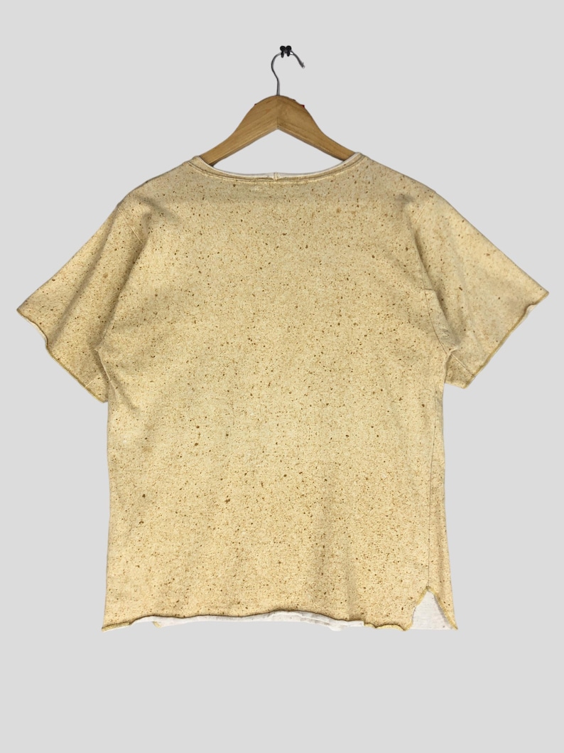May include: A short-sleeved, beige t-shirt with a speckled pattern, hanging on a wooden hanger. The shirt has a raw hemline and a slightly distressed appearance. The fabric appears to be a textured cotton blend.