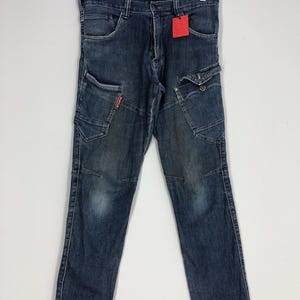 Puede incluir: Vaqueros de mezclilla azul oscuro con múltiples bolsillos y una etiqueta roja. Los vaqueros están colgados de una percha de madera. Los vaqueros tienen un diseño clásico de cinco bolsillos con bolsillos cargo adicionales en las piernas.