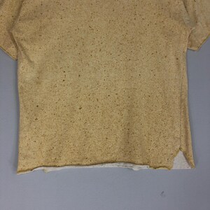 May include: A short-sleeved, tan-colored t-shirt with a speckled pattern. The shirt has a raw hemline and a split at the bottom hem. The fabric appears to be a soft, lightweight material.