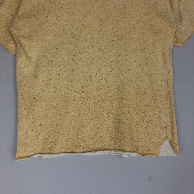 May include: A short-sleeved, tan-colored t-shirt with a speckled pattern. The shirt has a raw hemline and a split at the bottom hem. The fabric appears to be a soft, lightweight material.