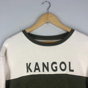 Może przedstawiać: Biała i oliwkowo-zielona bluza z nadrukiem słowa "KANGOL" dużymi, ciemnymi literami z przodu. Bluza ma okrągły dekolt i wisi na drewnianym wieszaku.