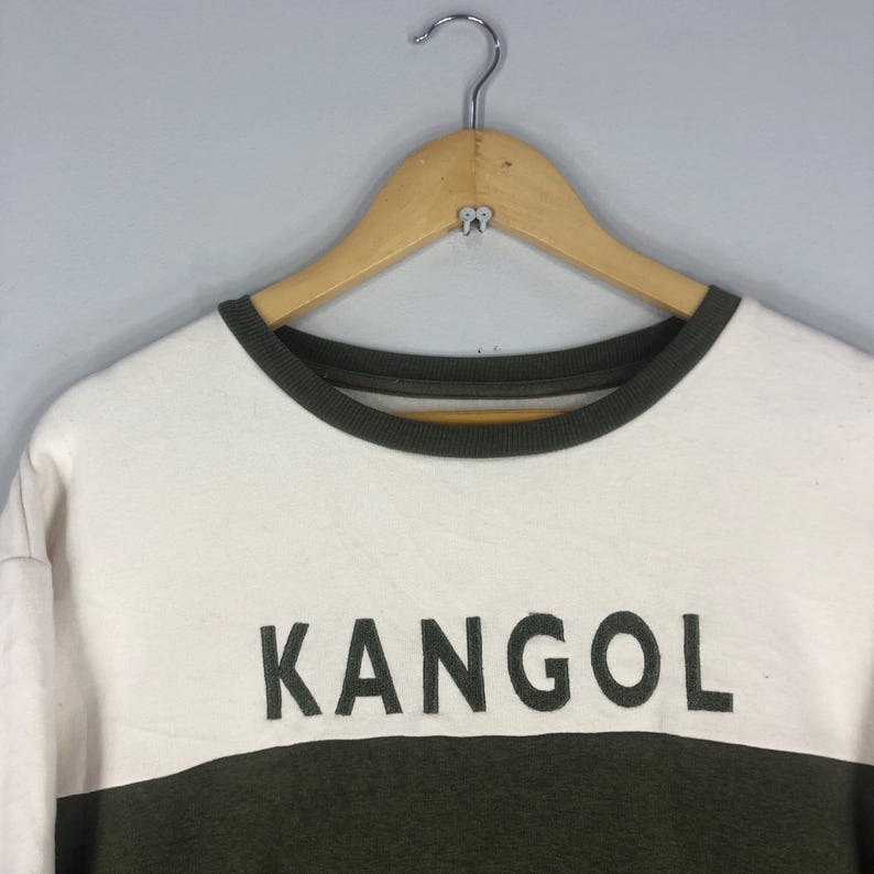 Może przedstawiać: Biała i oliwkowo-zielona bluza z nadrukiem słowa "KANGOL" dużymi, ciemnymi literami z przodu. Bluza ma okrągły dekolt i wisi na drewnianym wieszaku.