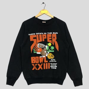 May include: A black long-sleeve sweatshirt featuring a graphic print that reads "SHOW DOWN IN THE SUN SUPER BOWL XXIII AFC CHAMPIONS JOE ROBBIE STADIUM MIAMI 1989" with an illustration of a football character wearing a helmet and sunglasses.