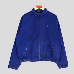May include: A royal blue zip-up jacket featuring a classic collar, long sleeves with elastic cuffs, and a ribbed hem. The garment displays a small red embroidered logo on the left chest and includes two front pockets with silver-toned snap closures.